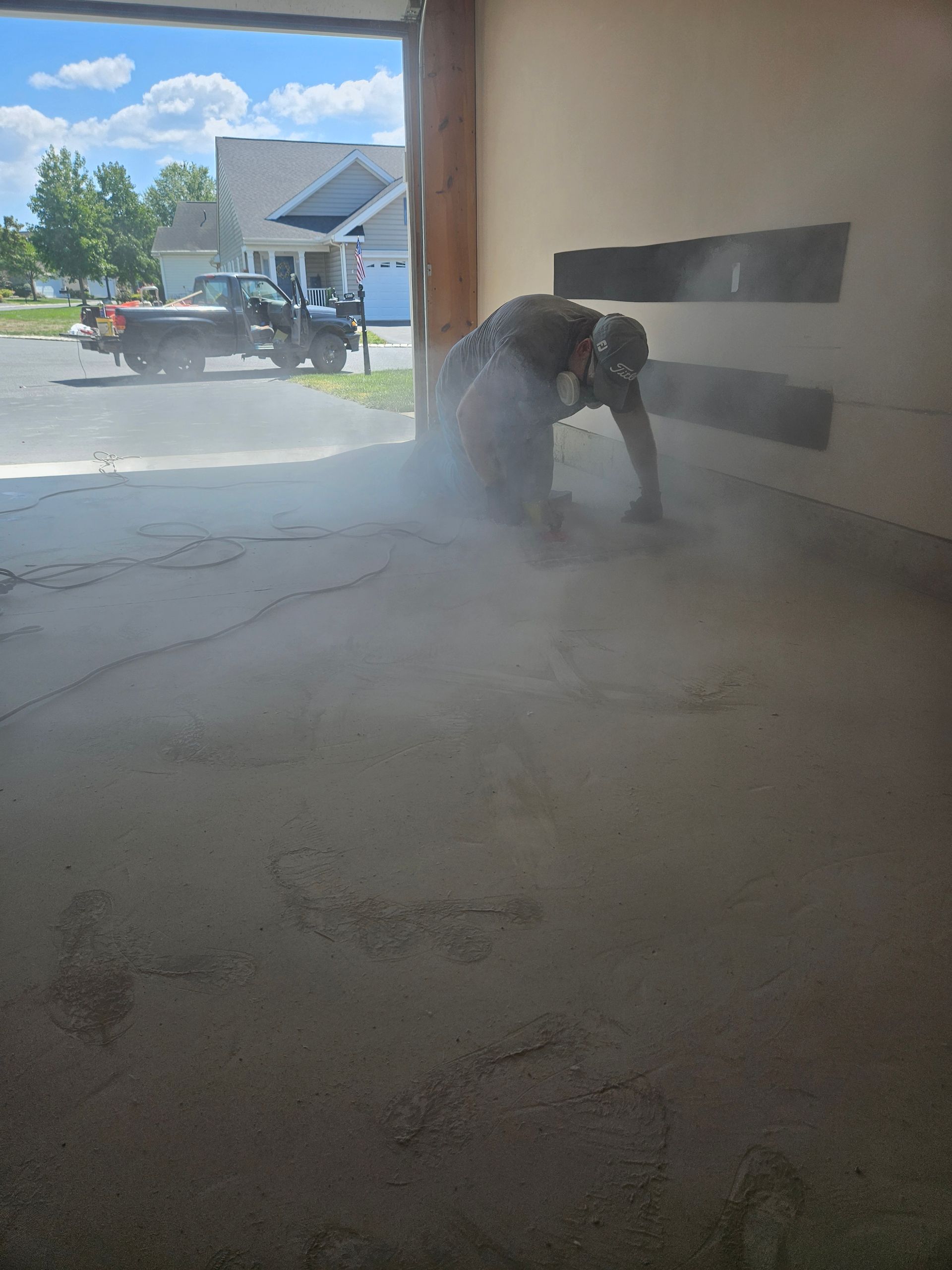 A person wearing a respirator grinds a concrete garage floor, creating a dense cloud of dust.