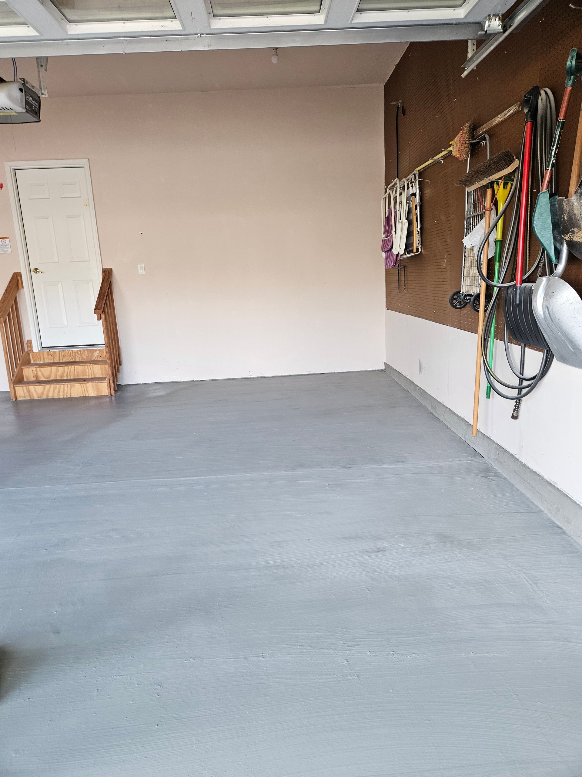 An empty garage with a gray painted floor, a doorway with wooden steps, and various tools hanging on the brown side wall.