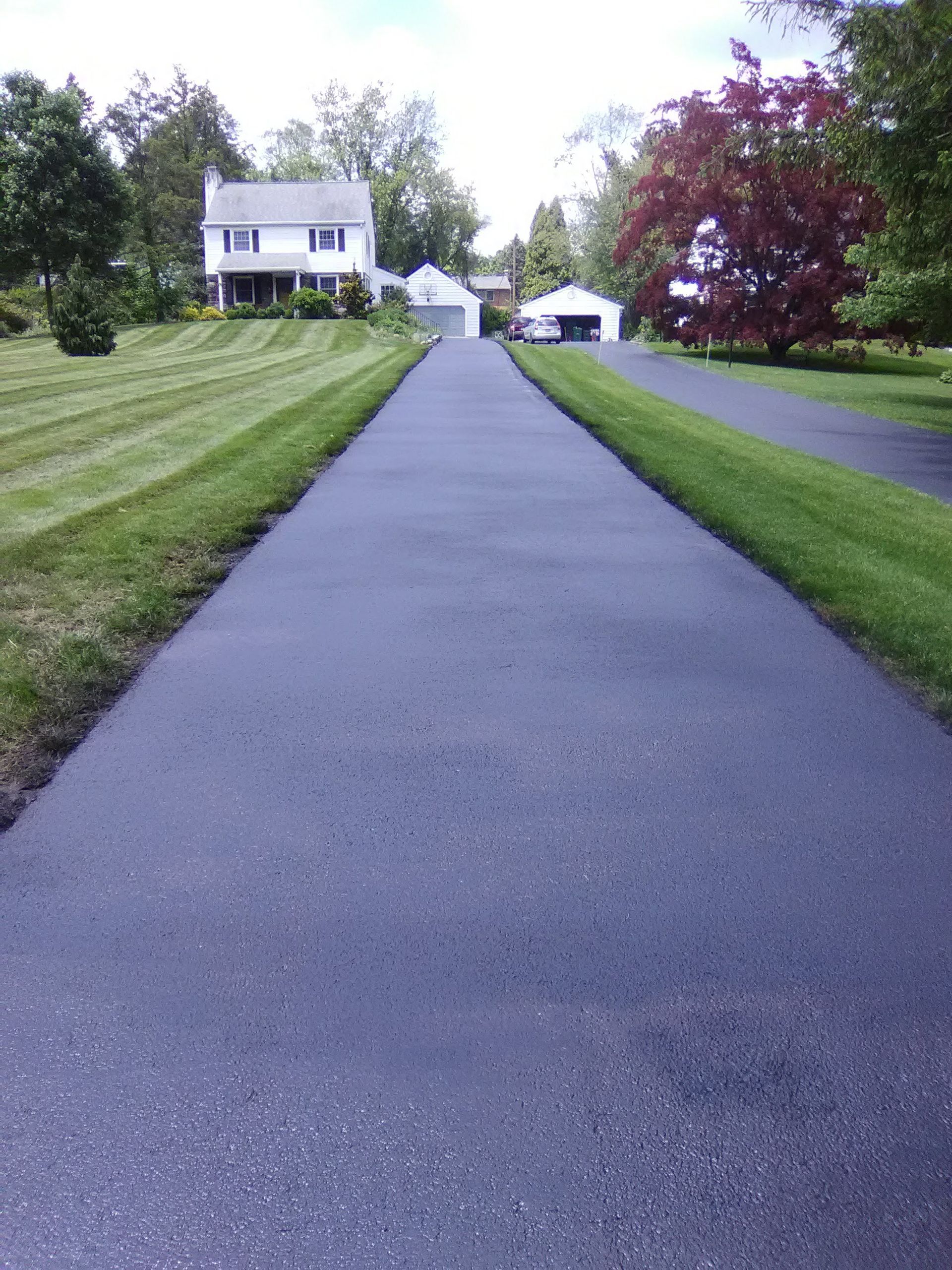 A paved asphalt driveway leads toward a white house with a garage in a suburban setting with green lawns and trees.