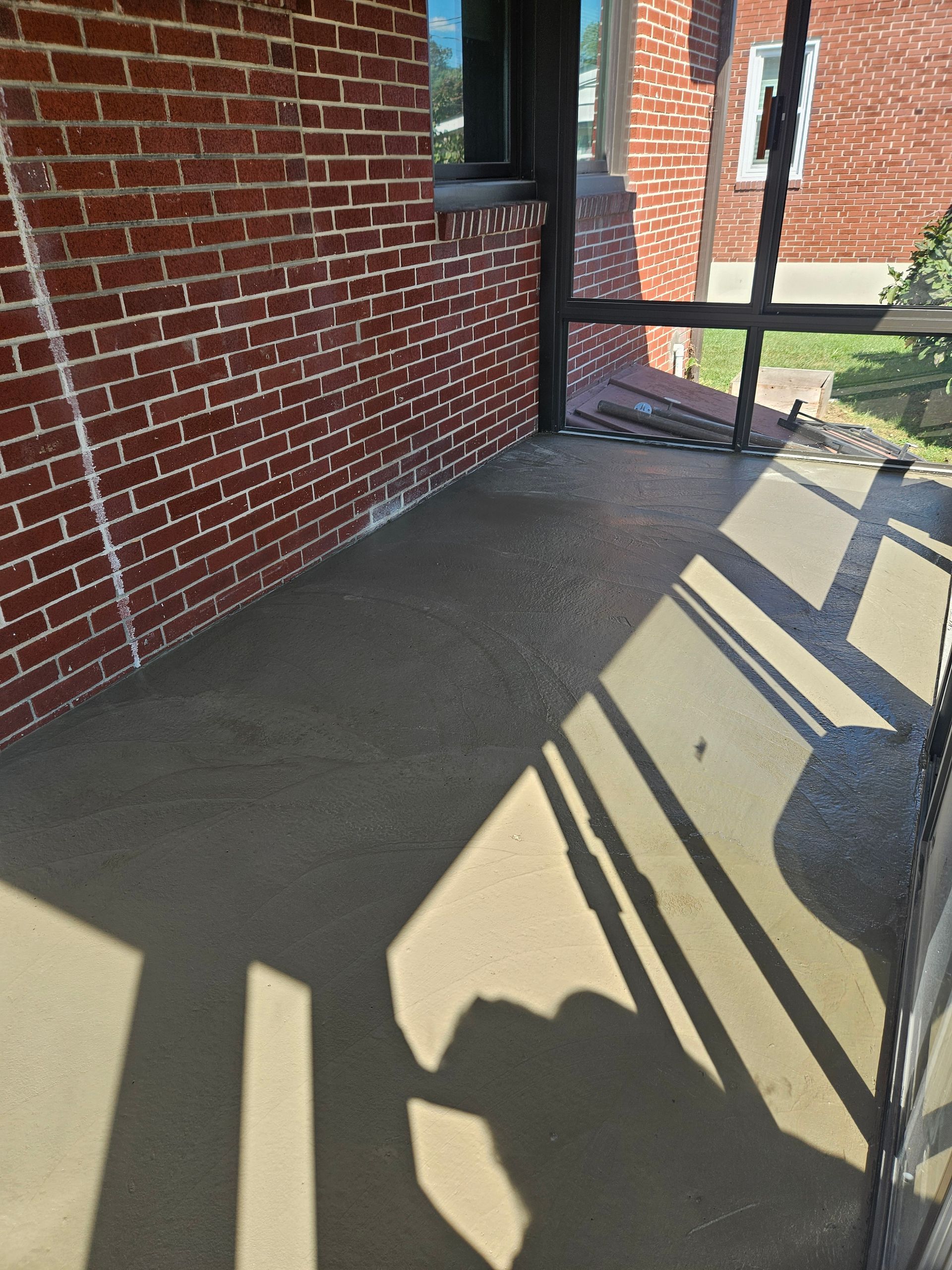 A concrete porch floor next to a red brick wall, with dark shadows cast by a screened-in enclosure.