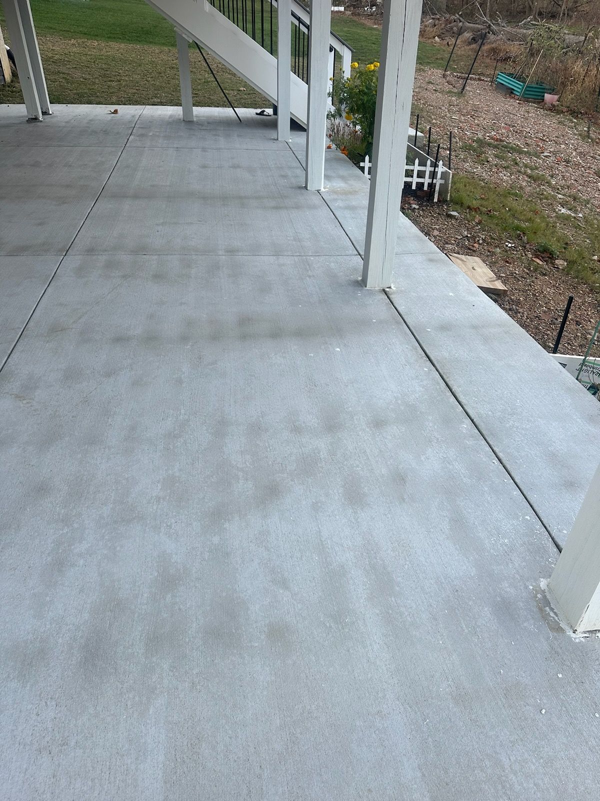 Concrete patio surface under a wooden deck, showing white support posts and a partial view of the stairs above.