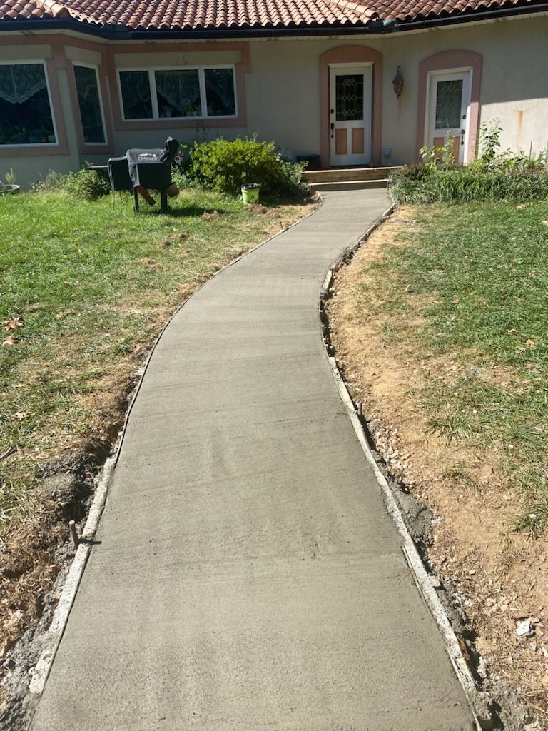A new, gray concrete walkway curves through a grassy lawn leading to the front entrance of a stucco house.