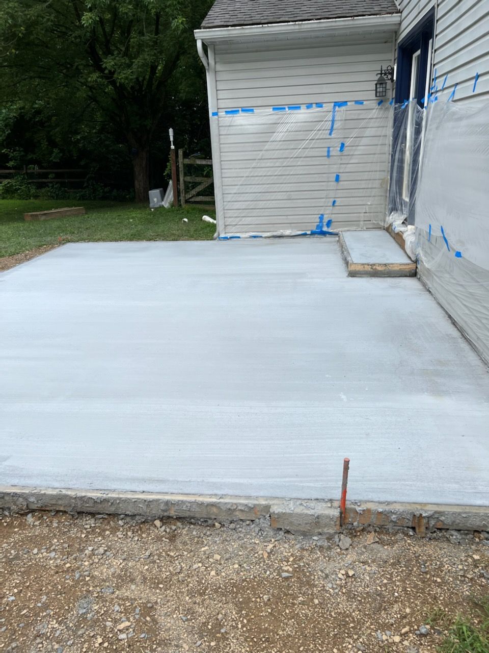 A newly poured concrete patio adjacent to a house with light gray siding and plastic protective sheeting.