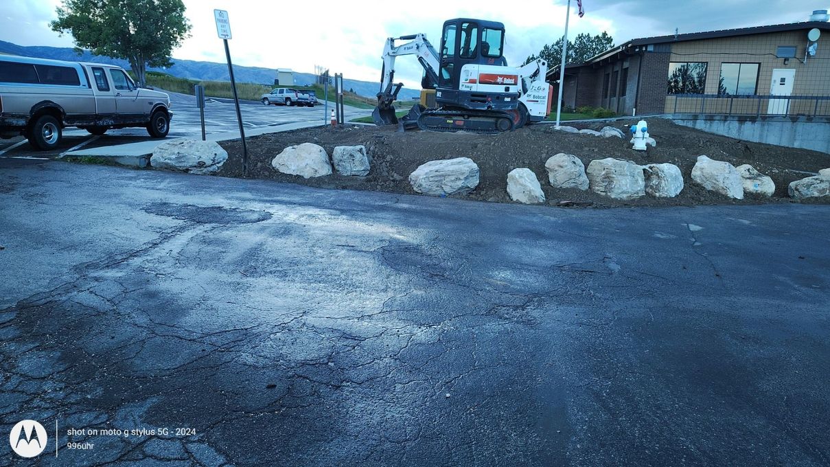 A construction excavator sits on a dirt mound bordered by large landscape stones in front of a building and parking lot.