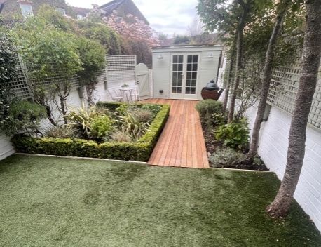 Neatly landscaped garden with artificial grass, central wooden walkway, shrubs, trees, and a small white shed.