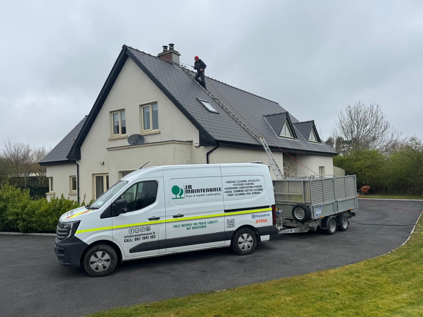 White service van and trailer parked beside a beige house with workers on the roof under cloudy skies