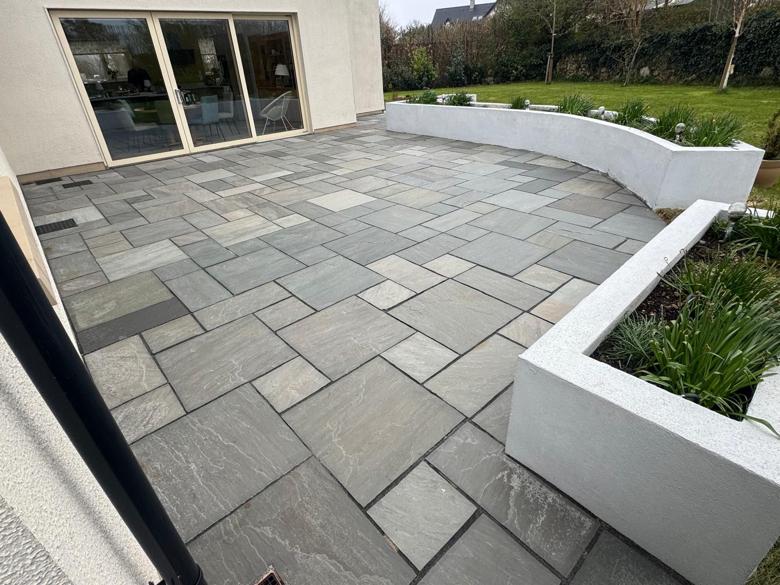 Modern patio with gray stone pavers, curved white planter beds, and sliding glass doors beside a building