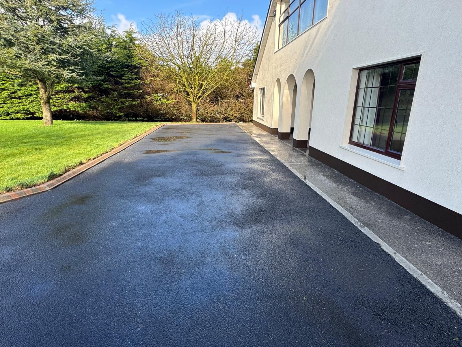 Wet driveway beside a white house, bordered by green lawn and trees on a sunny day