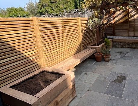 Sunlit patio with wooden fencing, raised planter, slate paving, and potted plants