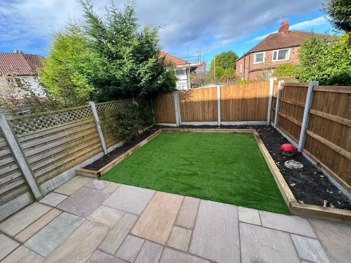 Small backyard patio with artificial grass, paving stones, wooden fences, and a hedge under a blue sky
