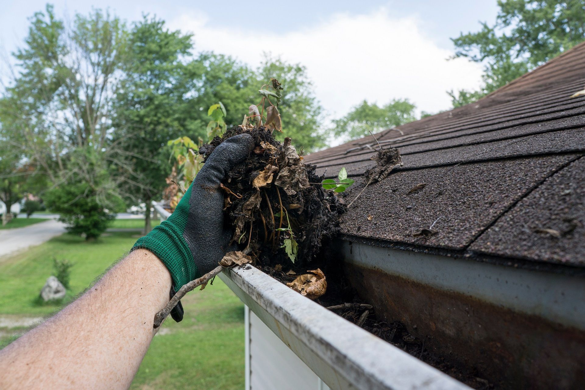 Gloved hand removing wet debris from a gutter beside a shingled roof