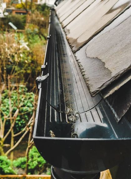 Black roof gutter filled with debris and leaves beside weathered shingles