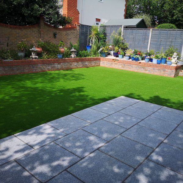 Sunny backyard with green lawn, gray patio tiles, and flower beds along a brick wall.
