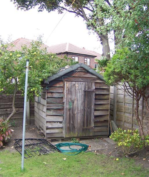 Weathered wooden garden shed beside a fence and trees in a backyard.