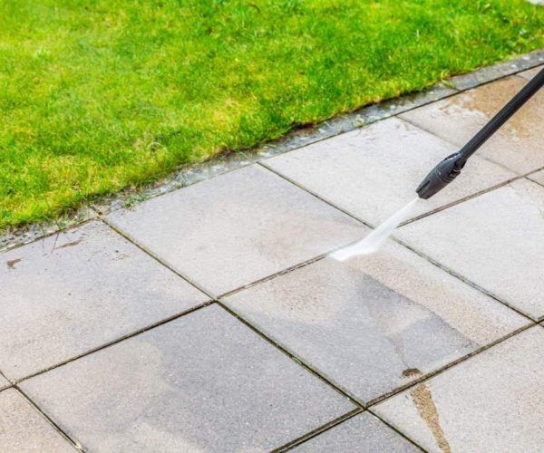 Pressure washing patio tiles beside a green lawn with a spray wand and wet streaks