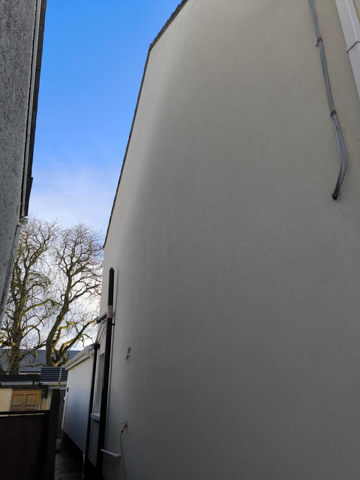 Narrow alley between white building walls under a clear blue sky, with trees and parked vehicles in the distance