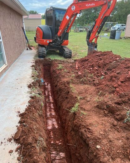 An excavator is digging a hole in the dirt in front of a house.