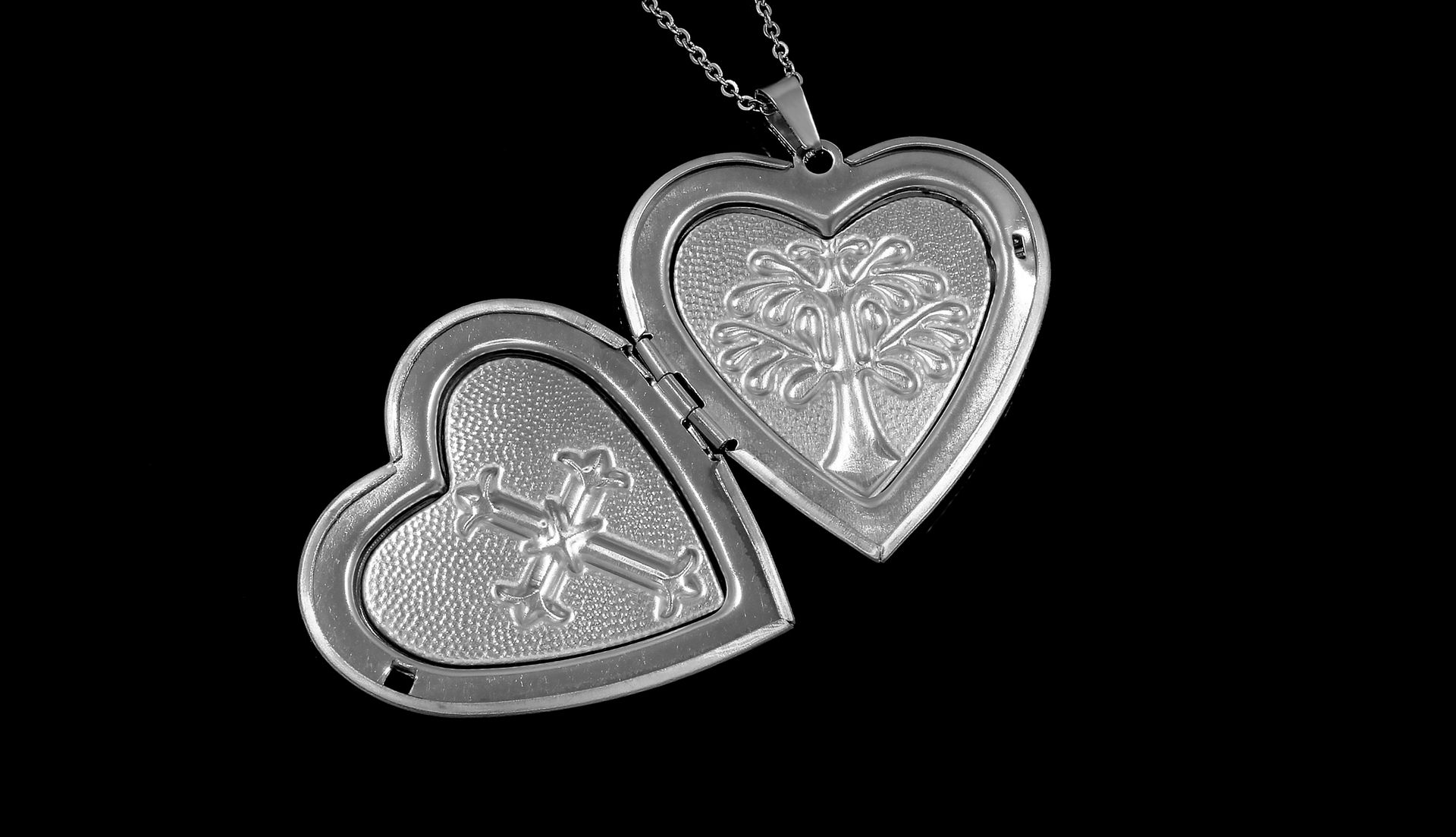 An open silver heart-shaped locket on a chain, showing intricate tree and cross designs inside on a black background.