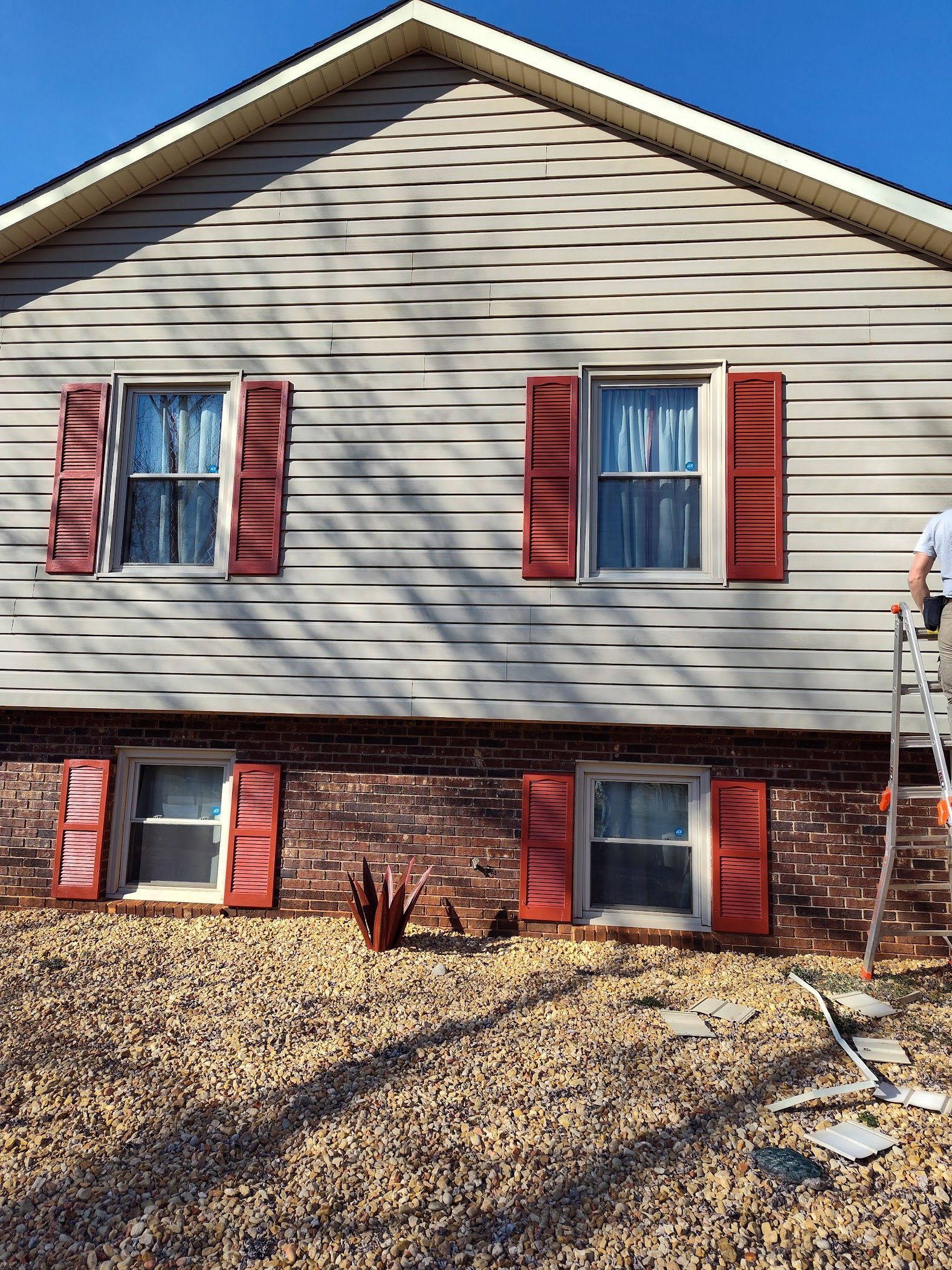 A man is standing on a ladder in front of a house with red shutters.