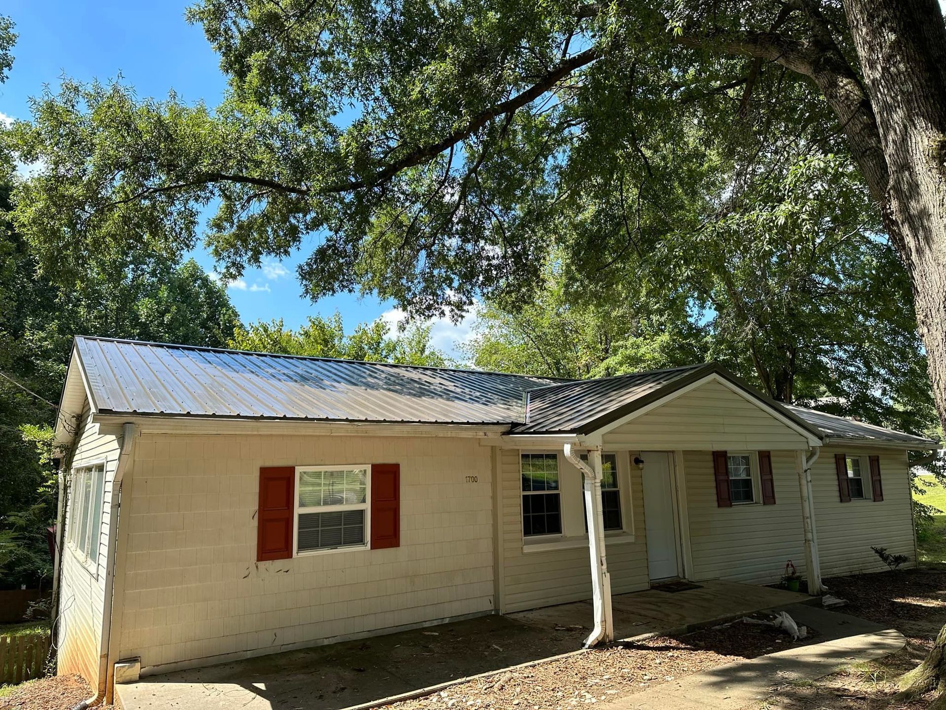 A white house with red shutters and a metal roof is surrounded by trees.