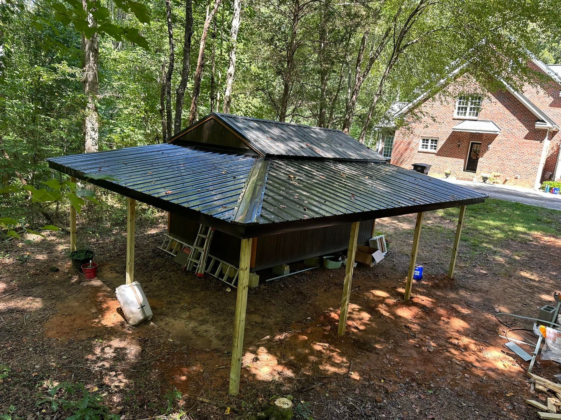 A shed with a metal roof is sitting under a canopy in the woods.