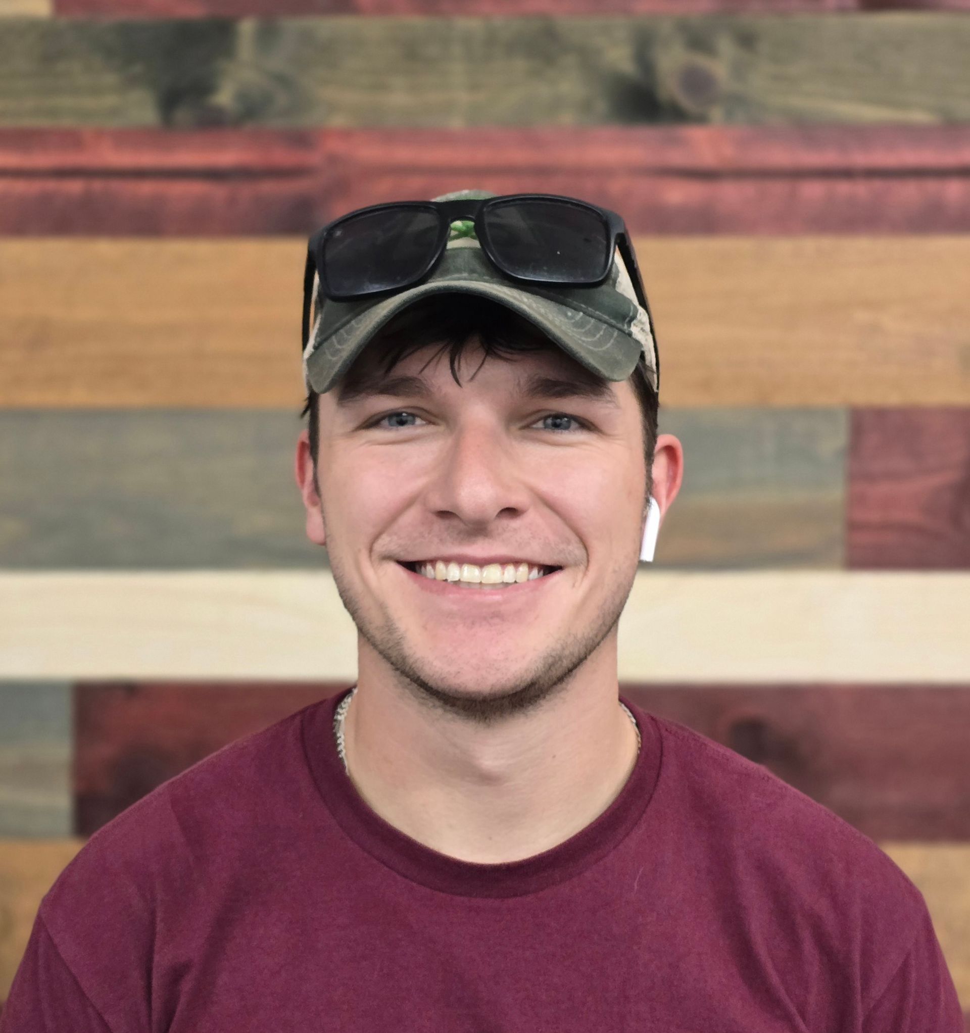 Smiling young man in cap, sunglasses, and maroon shirt, against a wooden background.