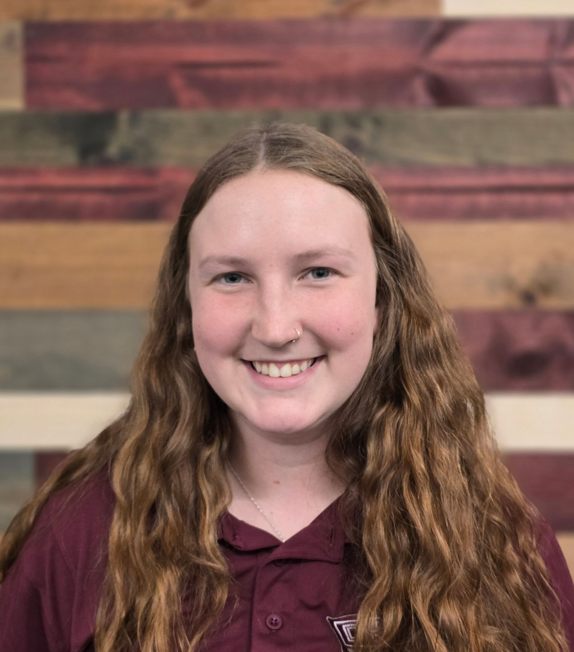 Young woman with long brown hair, smiling, wearing a maroon shirt, wooden background.
