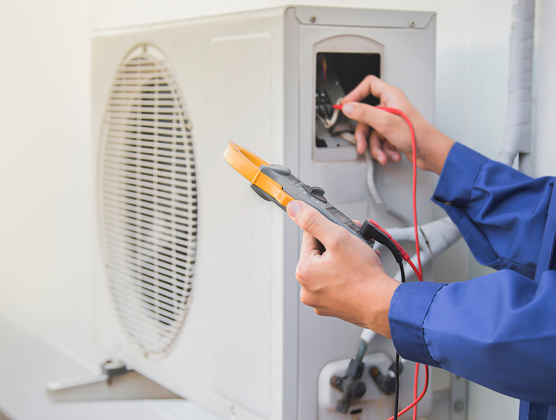 Technician in blue uniform tests an air conditioner's electrical components with a multimeter outdoors.
