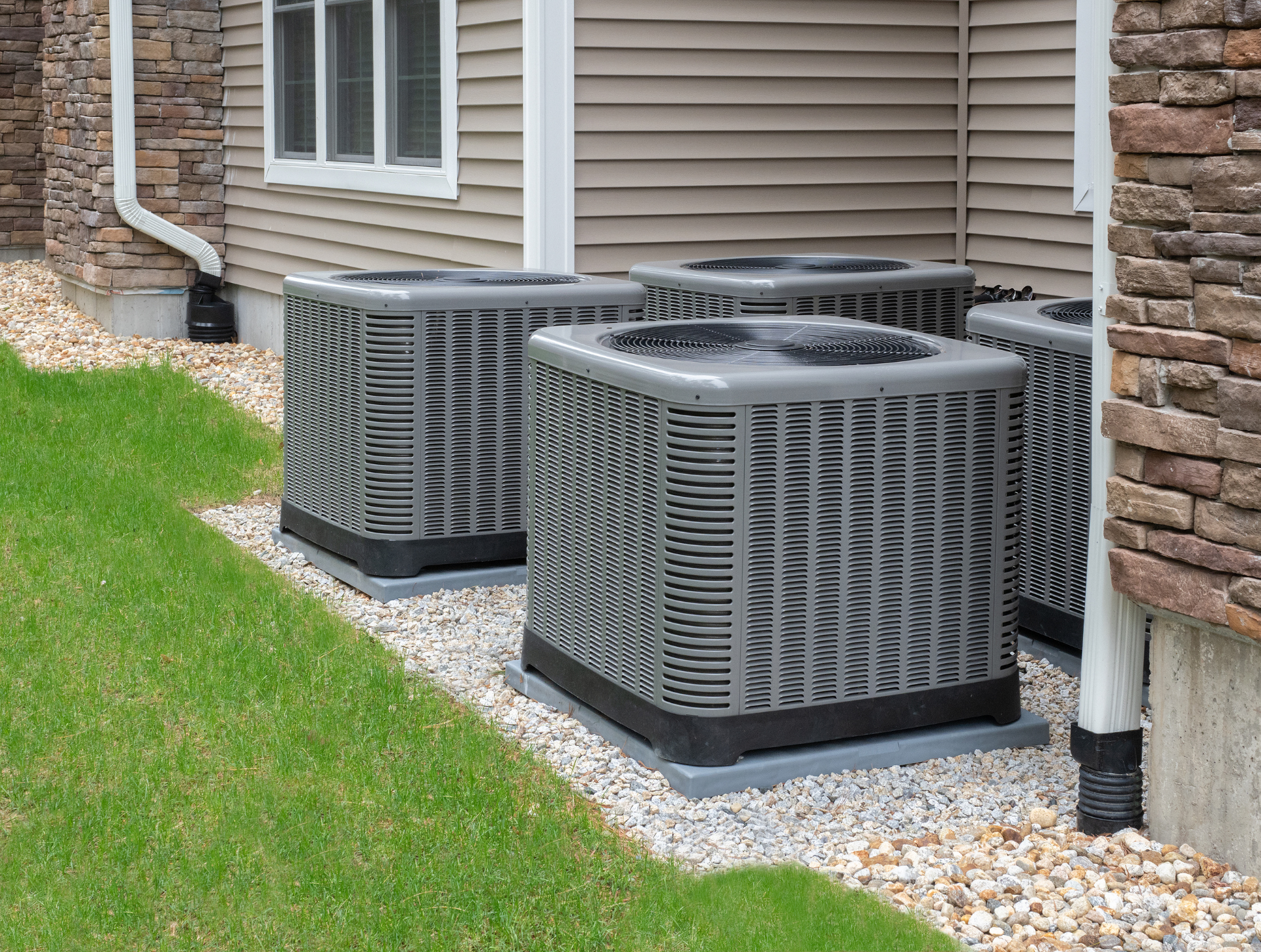 Four gray air conditioning units next to a building with a stone and beige exterior.