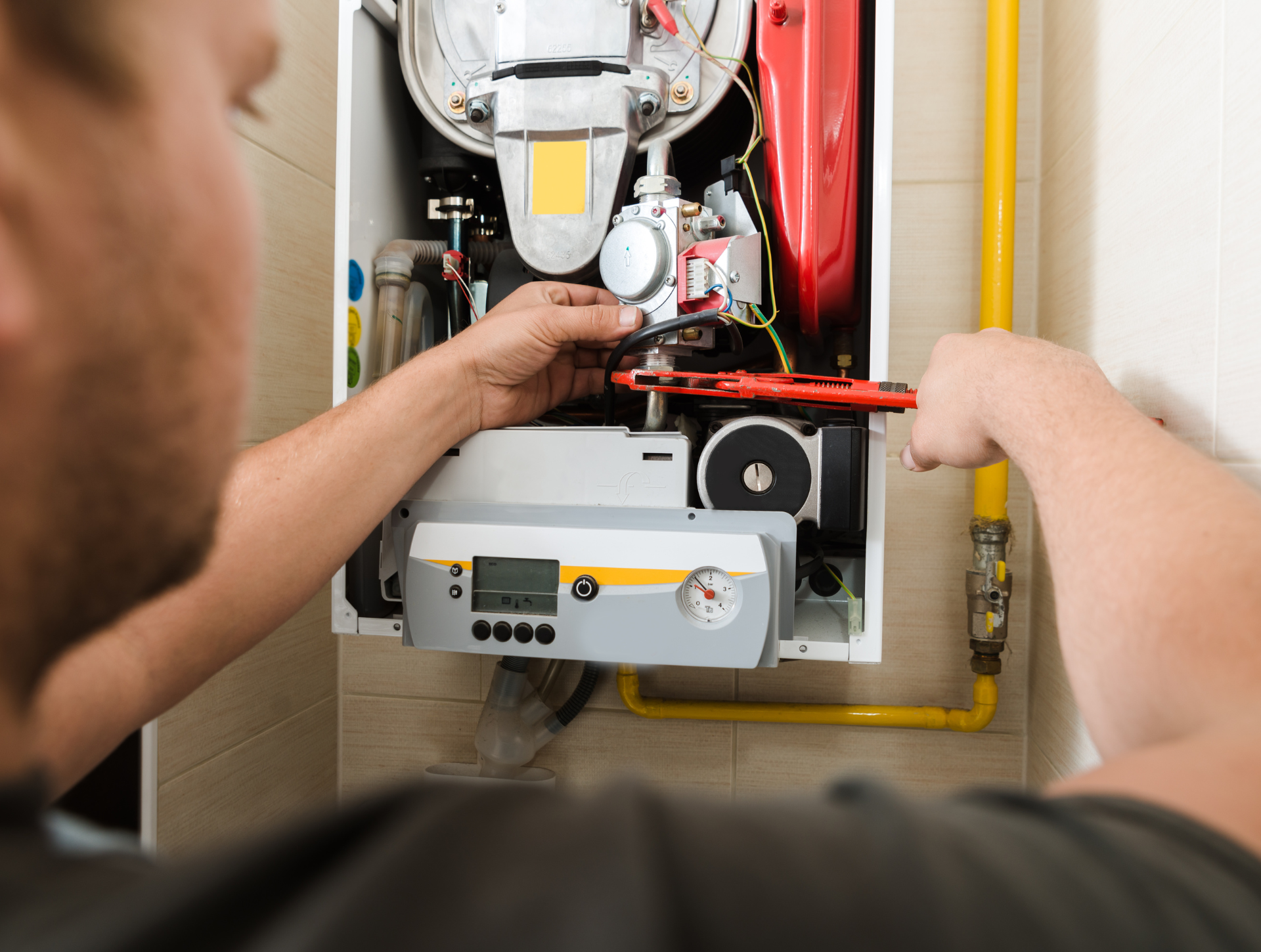 A person repairs a white wall-mounted furnace with a wrench, near yellow gas pipes.