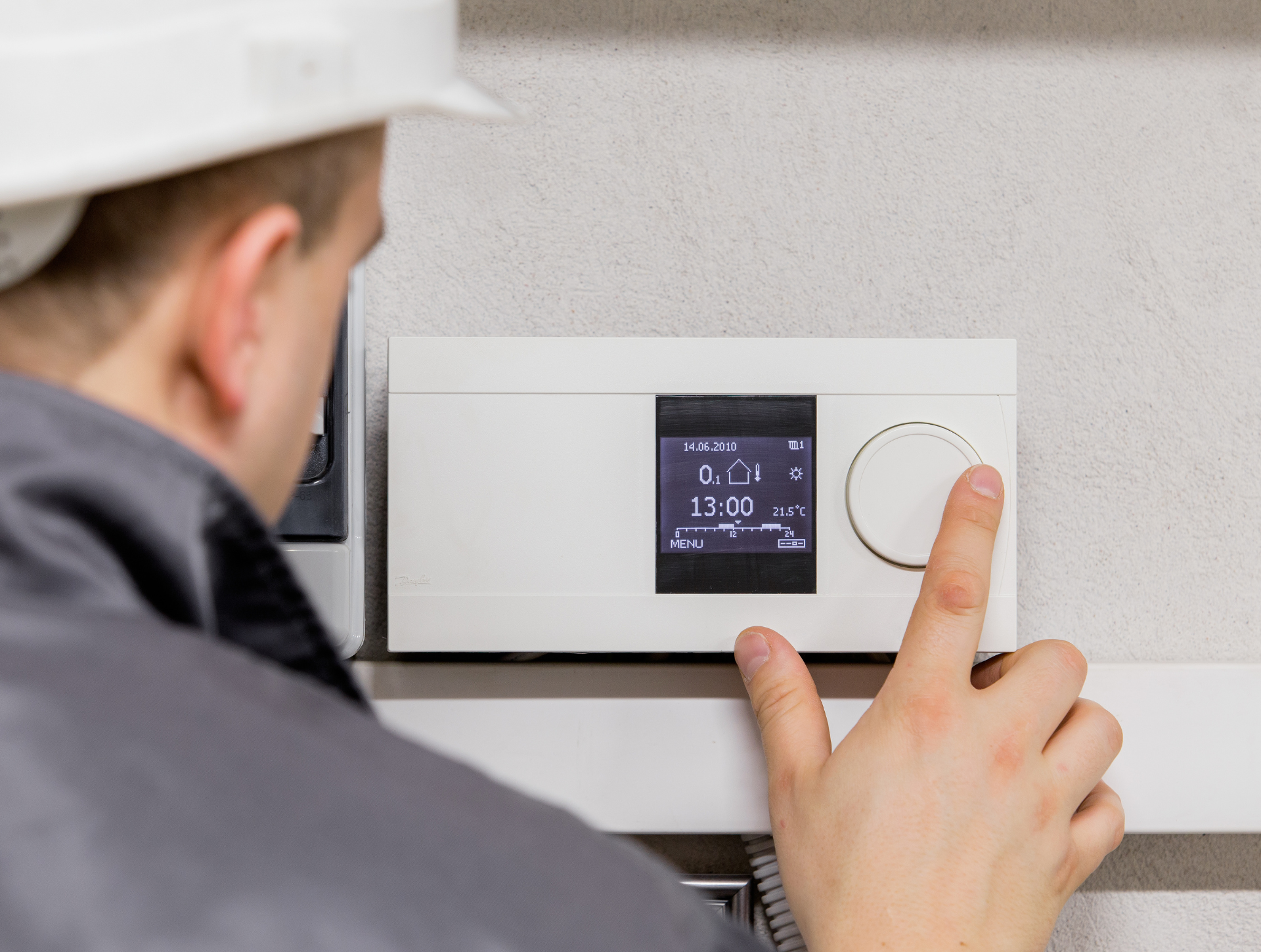Person in hard hat adjusting a white thermostat with a dial; indoors on a gray wall.