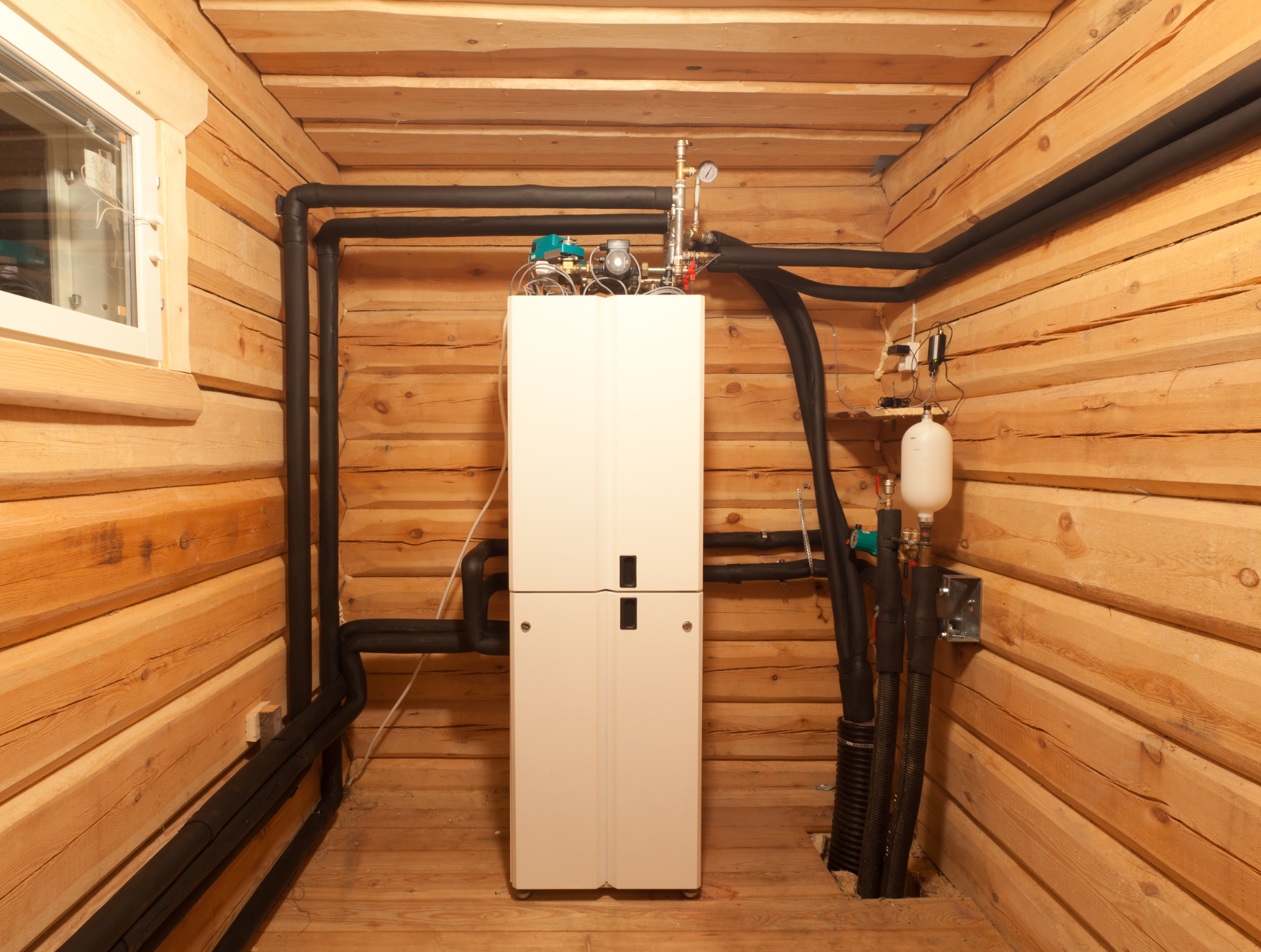 White heating system cabinet against wooden wall, with black insulated pipes.