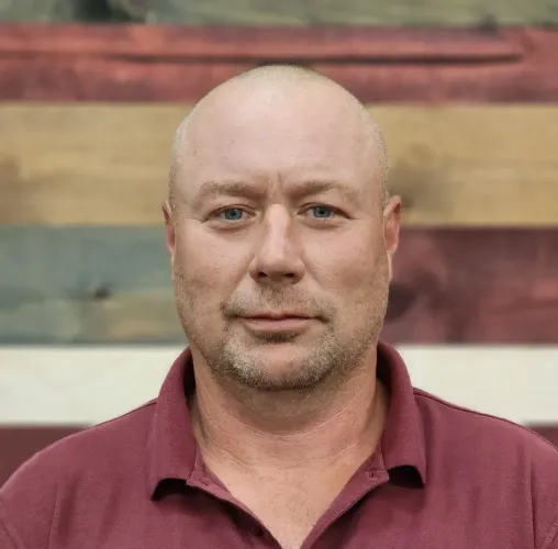 Bald man wearing a maroon polo shirt, looking at the camera, in front of a wooden wall.