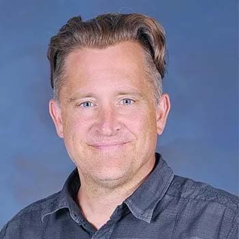 Man with short brown hair smiles, wearing a dark blue button-up shirt against a blue backdrop.