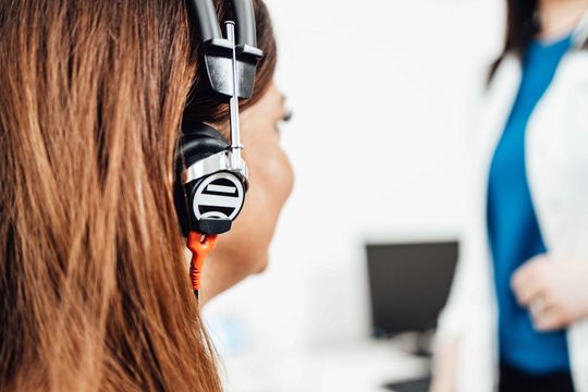 Woman wearing headset, facing away, in office setting. Another person in background.