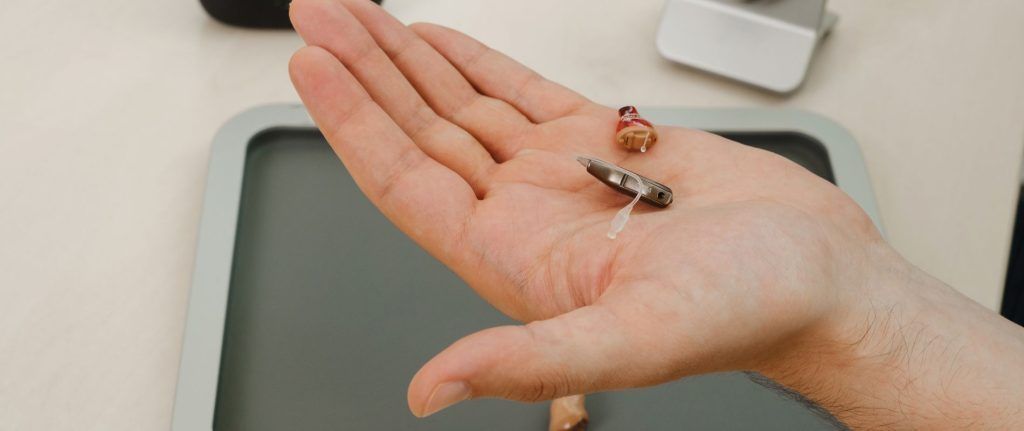 A person's open palm holding two hearing aids. A light grey tray is visible in the background.