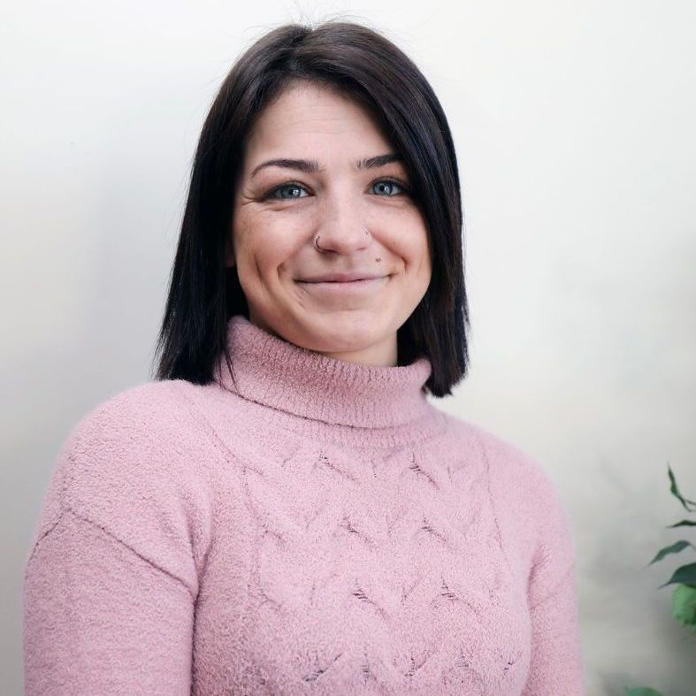 Woman with dark hair smiles, wearing a pink turtleneck sweater, in front of a neutral-colored wall.