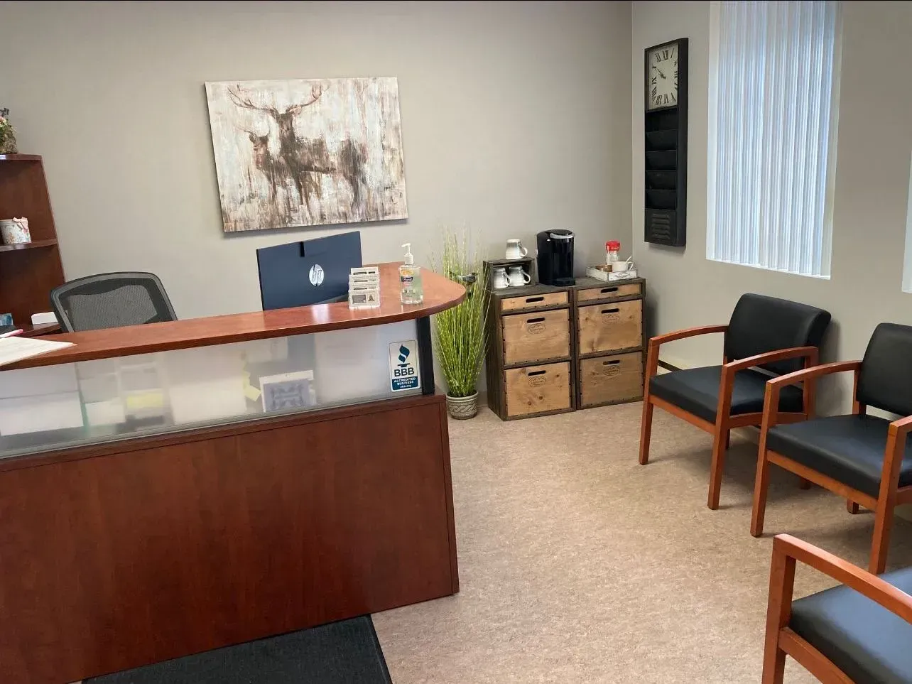 Office reception area with dark wood desk, two black chairs, artwork, and a coffee station.