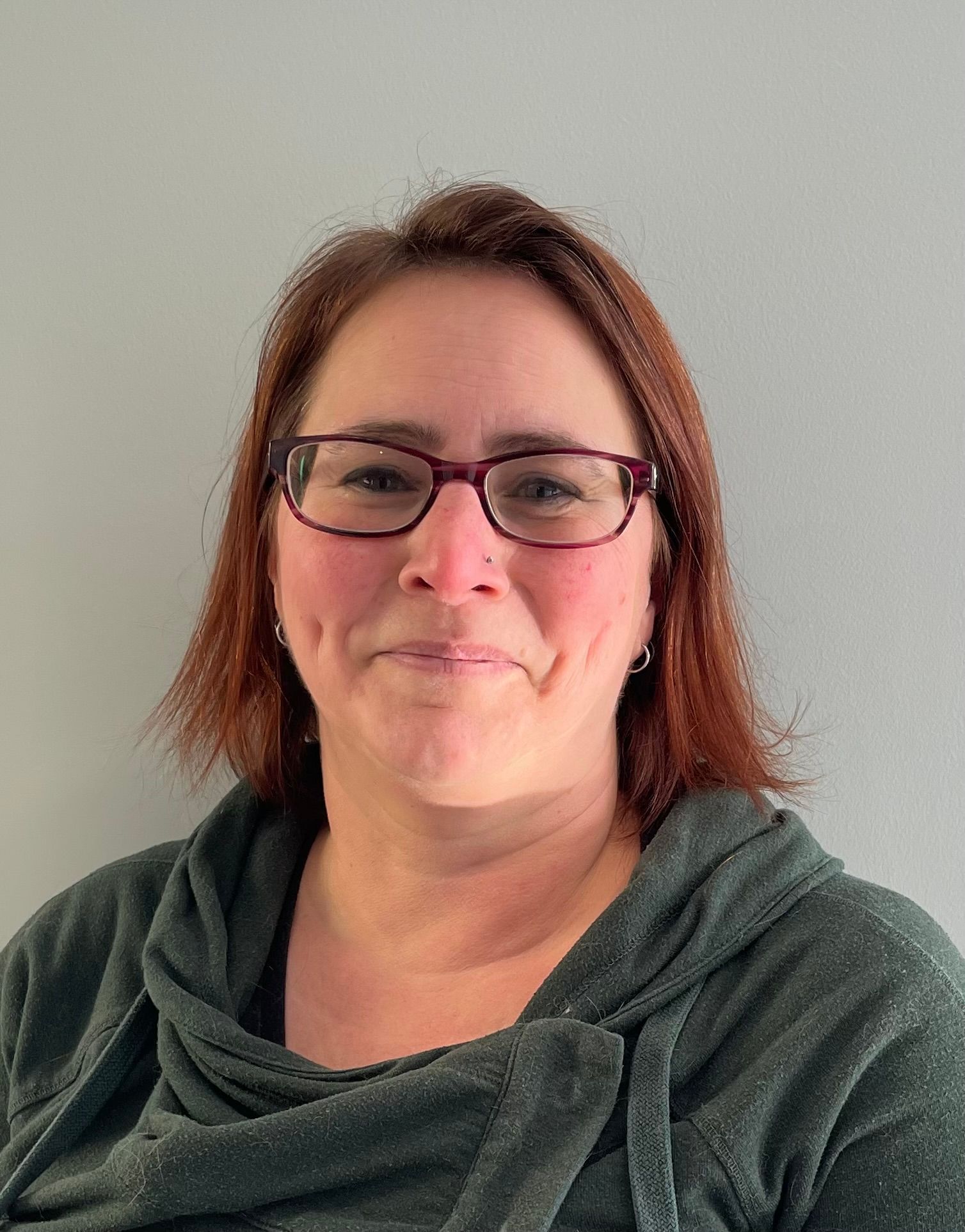 Woman with auburn hair and glasses wearing a green hooded sweatshirt smiles. Gray background.