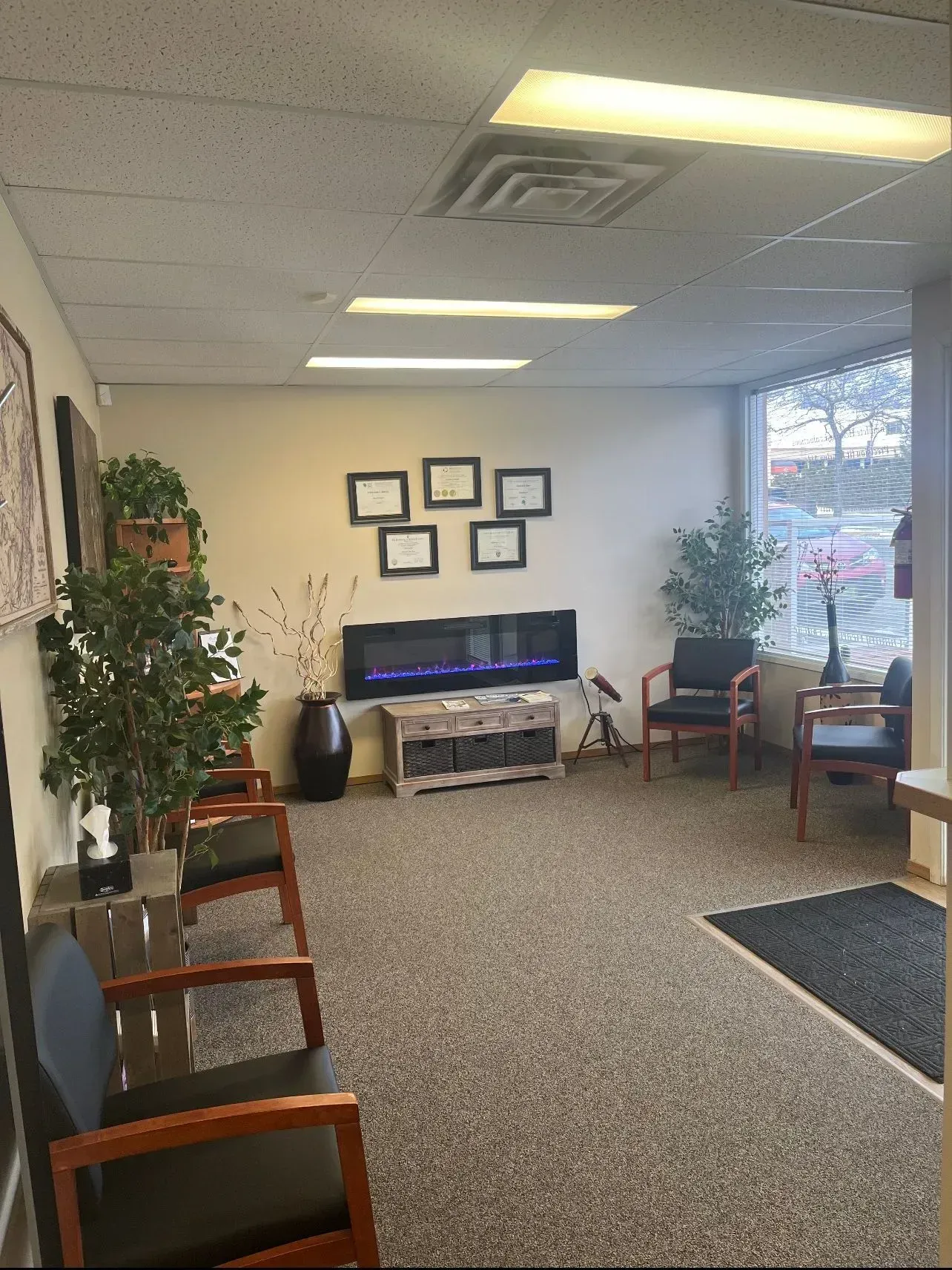 Waiting room with chairs, plants, fireplace, and framed documents on the wall.