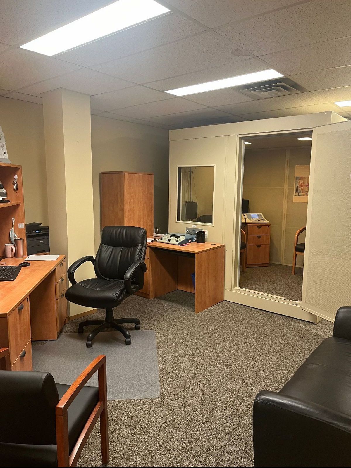Office interior with desks, chairs, and soundproof booth. Brown carpet and neutral walls, with overhead lights.