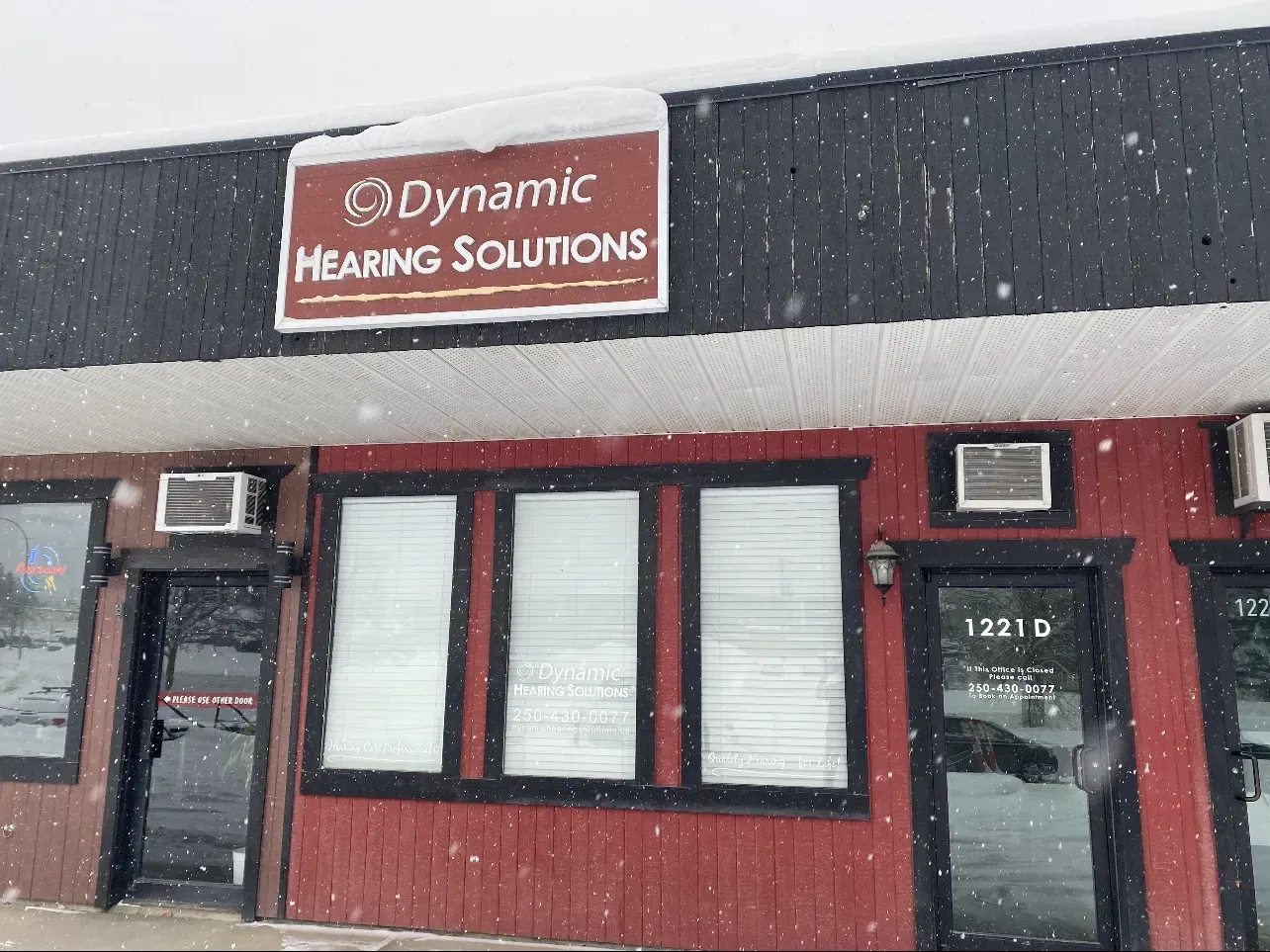 Building with sign for Dynamic Hearing Solutions, under snowfall. Red and black exterior, windows with blinds.