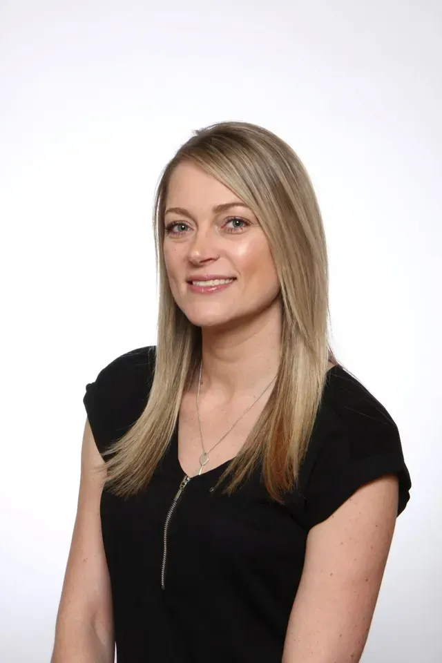 Woman with curly brown hair smiling, wearing a gray top, with a blue background.