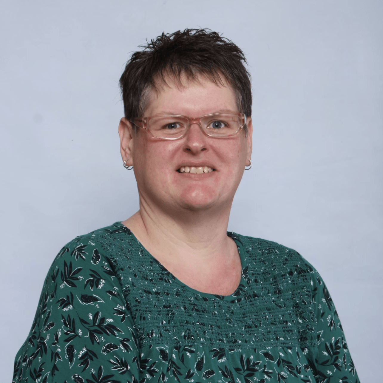 Woman with short brown hair, wearing glasses and a green patterned shirt, smiles.