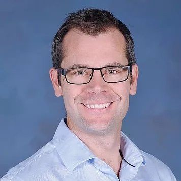 Man wearing glasses, smiling, blue button-down shirt, blue background.