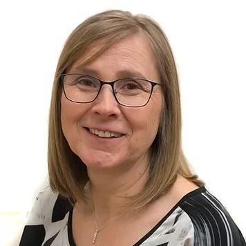 Woman with glasses smiles; wearing a black, white, and gray patterned top; neutral background.