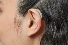 Close-up of a person's ear with a behind-the-ear hearing aid; gray hair.