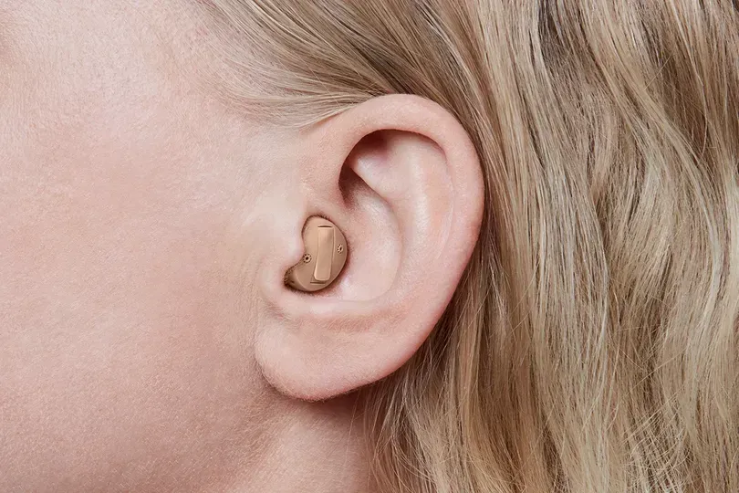 Close-up of a person's ear with a beige, in-the-ear hearing aid. Blonde hair frames the ear.
