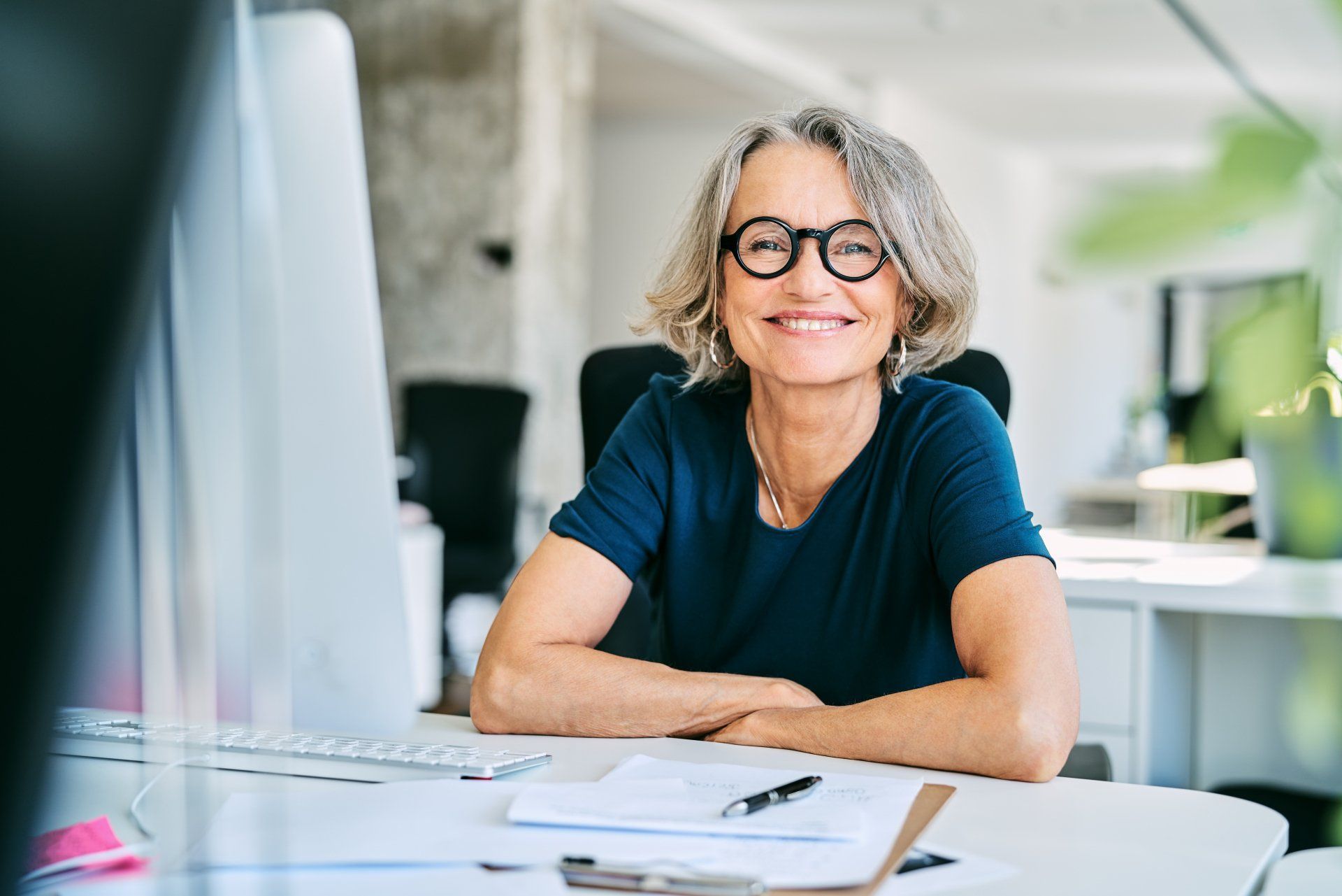 Woman with gray hair and glasses smiles at her desk, arms crossed.
