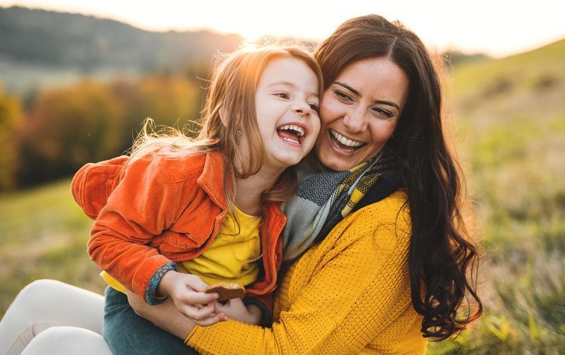 Woman and child laughing outdoors, embracing in a grassy field. Bright yellow and orange clothing.